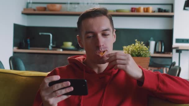 Portret van een man in rode hoodie die chips eet en video of film kijkt op zijn smartphone op de bank thuis — Stockvideo
