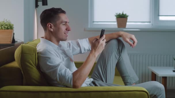 Joven sosteniendo dispositivo de teléfono inteligente mensajes de texto SMS mensaje sentado en el sofá en casa, sonriendo macho usando aplicaciones en el chat móvil en las redes sociales navegar por la web en el teléfono en el interior — Vídeo de stock