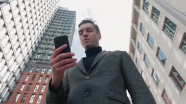 Şehir arka planında akıllı telefondan iş uygulaması kullanan bir adam. Akıllı telefondan iletişim kuran yakışıklı genç işadamı modern bina geçmişine güvenerek gülümsüyor..