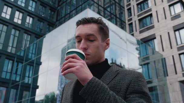 Portrait of ambitious businessman stand in the city center street and drinking coffee. Motivated young entrepreneur dreams of future achievements. Modern glass building on background. — Stock Video
