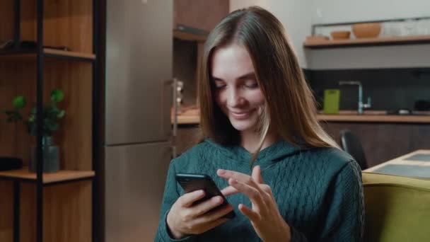 Gelukkig jonge vrouw houden smartphone kijken naar het scherm genieten met behulp van mobiele apps plezier spelen van games chatten in sociale media zitten op de bank thuis levensstijl — Stockvideo