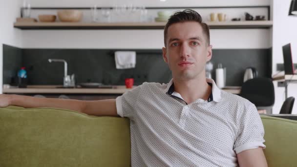 Portrait de l'homme heureux se repose tout en étant assis sur le canapé à la maison. Un jeune homme aime le style de vie en regardant la caméra assis sur un canapé confortable dans son appartement. — Video