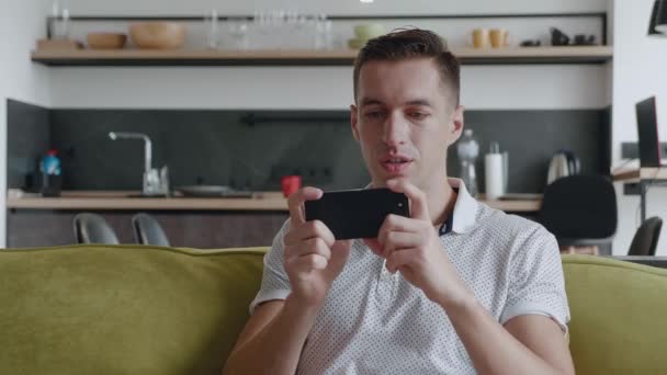 Akıllı telefonuyla oynayan genç adam heyecan verici mobil uygulaması evdeki oturma odasında kanepede otururken yumruğunu sıkarak bir yarış oyunu kazanıyor.. — Stok video