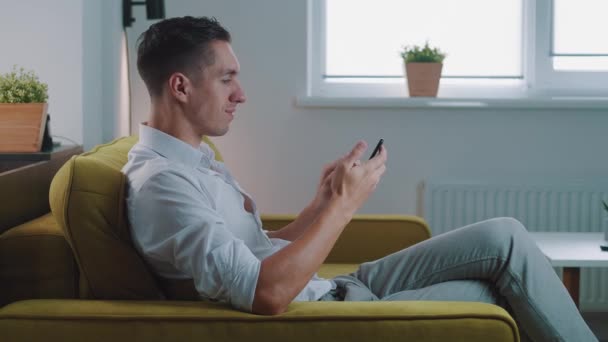 Jovem segurando dispositivo de smartphone mensagem SMS sms sentado no sofá em casa, sorridente masculino usando aplicativos em bate-papo móvel em redes sociais navegar na web no telefone dentro de casa — Vídeo de Stock