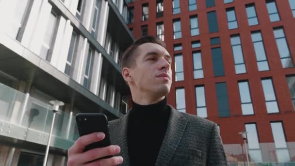 Portrait d'homme d'affaires ambitieux debout dans le centre-ville et en utilisant smartphone. Les jeunes entrepreneurs rêvent de réalisations futures. Bâtiment moderne sur fond. — Video