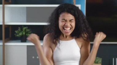 Evet, kazanan jest. Başarı kutlaması yapan Afro-Amerikan genç bayan, kameraya şaşırdı, evinde oturuyor. Zafer konsepti.