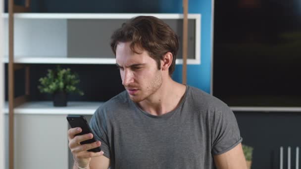 Homem chocado lendo notícias terríveis em seu smartphone em casa apartamento fundo. Desagradavelmente surpreendido jovem macho reagindo mensagem em seu telefone celular. — Vídeo de Stock