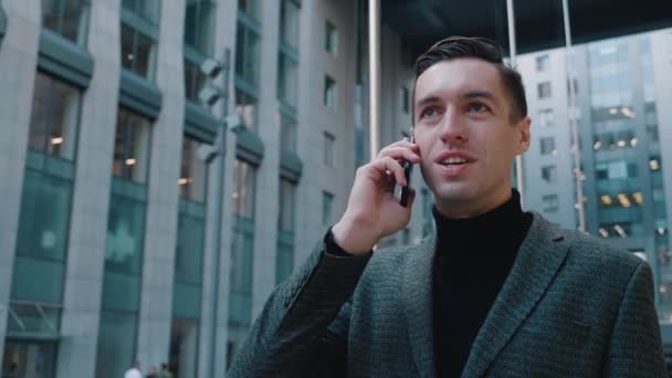 L'uomo d'affari sorridente di successo sta parlando su smartphone all'aperto vicino a edificio moderno. Il ragazzo e 'nel quartiere degli affari e parla al cellulare. Sembri soddisfatto. Stile di vita aziendale. — Video Stock