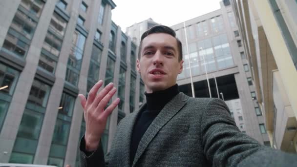 ビジネス街の屋外に立って自撮りビデオ通話をする若いビジネスマンを笑顔。現代のオフィスビルの近くに屋外でビデオブログを撮影ビジネスマンブロガーの肖像. — ストック動画