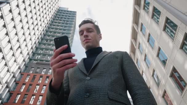 Muž používající obchodní aplikaci na chytrém telefonu na pozadí města. Fešák mladý podnikatel komunikuje na smartphone s úsměvem jistý na moderním pozadí budovy.