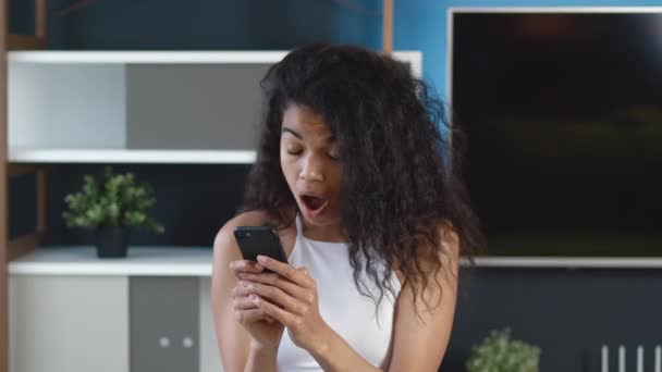 Εκπλήσσομαι που μια Αφροαμερικανή κοιτάζει το smartphone, αναρωτιέται και λέει ουάου. Σύγχρονο γραφείο ή σαλόνι φόντο. — Αρχείο Βίντεο