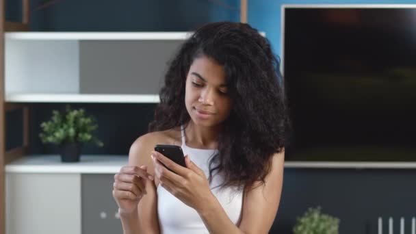 Νεαρή Αφρικανή θετική γυναίκα που χρησιμοποιεί smartphone surfing στα μέσα κοινωνικής δικτύωσης, ελέγχει ειδήσεις, στέλνει μηνύματα και χαμογελά. Χαριτωμένο χαρούμενο μαύρο κορίτσι με σγουρά μαλλιά χρησιμοποιώντας το κινητό τηλέφωνο στο σπίτι ή σύγχρονο γραφείο. — Αρχείο Βίντεο