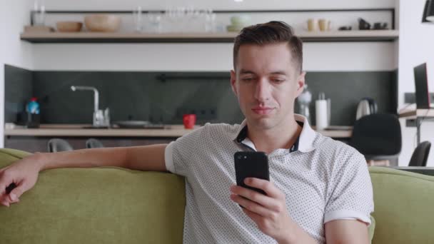 Šokovaný muž Reagující zpráva na Smartphone si zakryje ústa hrůzou, když sedí doma na pohovce v obývacím pokoji. Nepříjemně překvapená a vyděšená. Muž je šokován tím, co viděl na svém. — Stock video