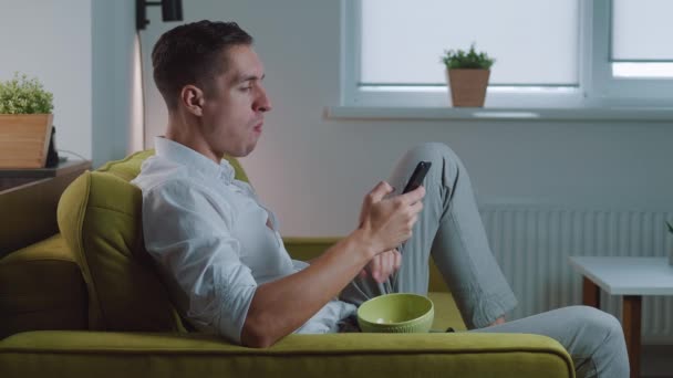 Retrato del hombre comiendo papas fritas y usando su teléfono inteligente mientras se relaja en el sofá en casa. Hombre sonriente comiendo bocadillos y usando aplicaciones en el chat móvil en las redes sociales mientras está sentado en el sofá. — Vídeos de Stock