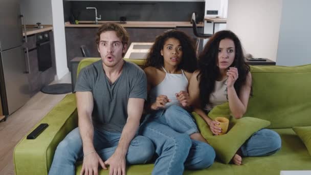 Група з трьох багатоетнічних друзів дивиться фільм жахів по телевізору, сидячи на дивані вдома. Молоді різноманітні люди розслабляються на дивані вдома під час перегляду трилерної плівки у вітальні — стокове відео