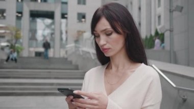 Kendine güvenen iş kadını, büyük modern iş merkezinin yakınındaki Smartphone 'da mesajlaşıyor. İş bölgesindeki cep telefonunda uygulamayı kullanan genç bir kadın..