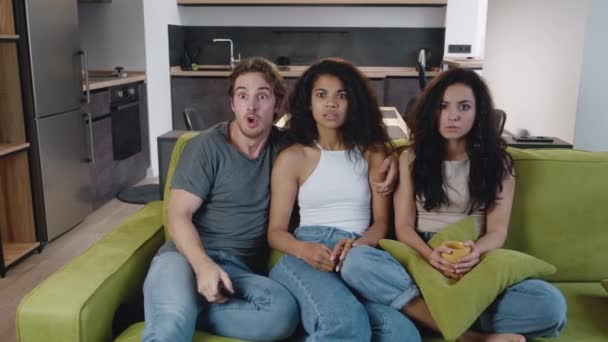 Grupa trzech wielonarodowych przyjaciół ogląda horror w telewizji, siedzi na kanapie w domu. Młodzi, zróżnicowani ludzie relaksujący się na kanapie w domu oglądając thriller w salonie — Wideo stockowe