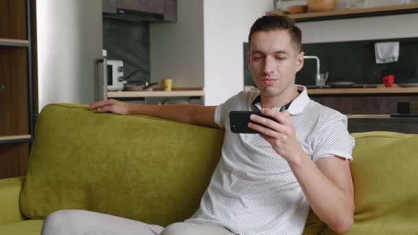 Κοντινό πλάνο της ανάπαυσης νεαρός άνδρας κάθεται στον καναπέ βλέποντας βίντεο στο smartphone στο σύγχρονο διαμέρισμα — Αρχείο Βίντεο