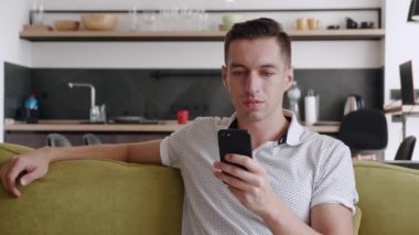 Modern apartmandaki akıllı telefon tebessümü kullanarak kanepede oturan neşeli genç adama yakın çekim teknolojisi mesajları yolluyorum.