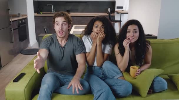 Divertido grupo joven de amigos multiétnicos celebran ganar la victoria del equipo de televisión objetivo juntos, ver el juego deportivo en casa y el apoyo. Un hombre y dos amigas ven fútbol en la televisión en casa en el sofá. — Vídeo de stock