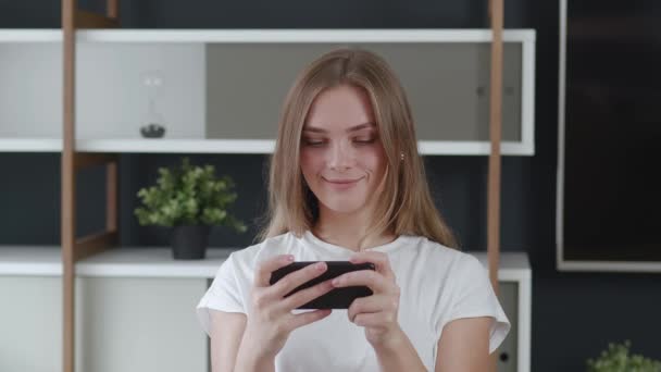 Sonriente joven mujer jugando juego de aplicación de teléfono celular en casa. Joven mujer caucásica disfrutando de los videojuegos smartphone. — Vídeo de stock