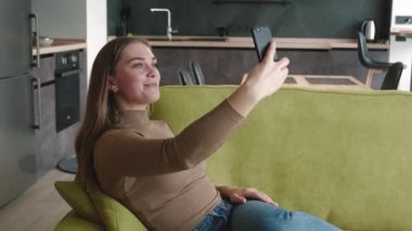 Evde akıllı telefon kullanarak arkadaşınla görüntülü sohbet eden mutlu bir genç kadın. Oturma odasındaki kanepede dinlenirken selfie çeken güzel bir kadın.