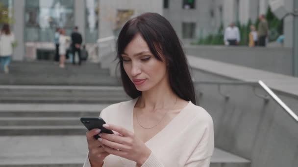 Šťastná pozitivní podnikatelka ženského šéfa s smartphonem textové zprávy, zatímco stojí venku ve městě obchodní čtvrti. Mladá usměvavá žena pomocí mobilní aplikace v blízkosti moderní kancelářské budovy. — Stock video