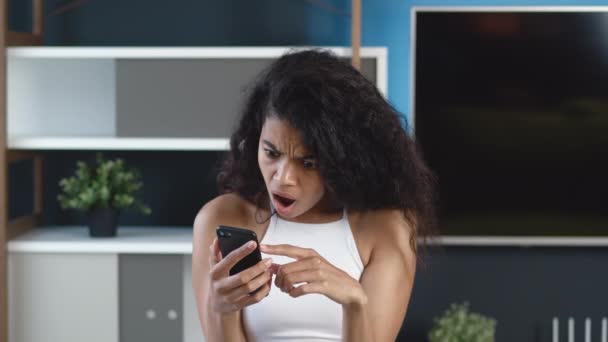 Шокированная африканская американка читает ужасные новости на своем смартфоне дома. Неприятно удивленная молодая женщина реагирует на сообщение на мобильный телефон. — стоковое видео