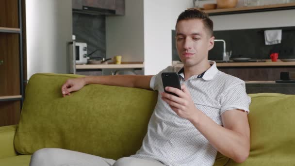 Κοντινό πλάνο του χαρούμενου νεαρού άνδρα που κάθεται στον καναπέ χρησιμοποιώντας το χαμόγελο smartphone στο σύγχρονο διαμέρισμα γραπτών μηνυμάτων κύλιση τεχνολογία υποκλοπή — Αρχείο Βίντεο