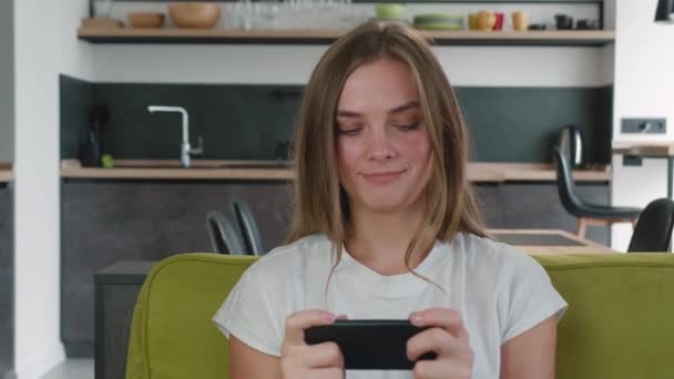 Lächelnde junge Frau beim Handy-App-Spiel im Wohnzimmer. Junge Kaukasierin genießt Smartphone-Videospiele, während sie im Wohnzimmer auf dem Sofa sitzt. — Stockvideo