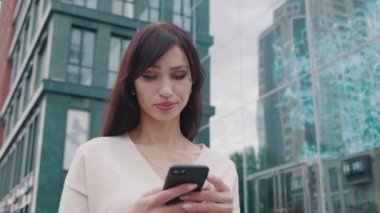 Kendine güvenen iş kadını, büyük modern iş merkezinin yakınındaki Smartphone 'da mesajlaşıyor. İş bölgesindeki cep telefonunda uygulamayı kullanan genç bir kadın..