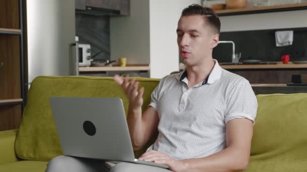 Hombre joven que utiliza el chat de vídeo en el ordenador portátil para comunicarse con amigos o socios de negocios. Hombre guapo está hablando y mirando a la pantalla del ordenador portátil en el sofá en casa sala de estar — Vídeos de Stock