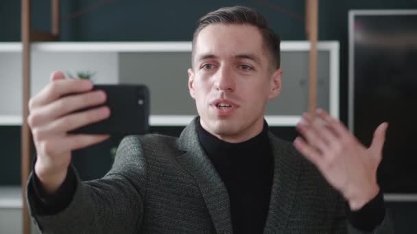Portrait of happy businessman making facetime video calling with smartphone at modern office, waving at people on phone screen. Young male in suit making selfie video chat online on mobile phone — Stock Video