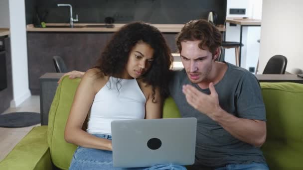 Geschokt gemengde etniciteit jong paar op zoek naar een laptop lezen van slecht nieuws tijdens het zitten op de bank thuis in de woonkamer. — Stockvideo