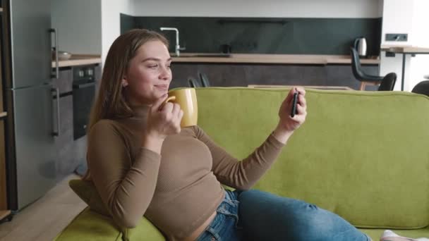 Улыбающаяся женщина наслаждается просмотром видео на смартфоне и пьет чай или кофе, лежащий на уютном диване в гостиной. Молодая женщина отдыхает на диване с горячим напитком и с помощью мобильного телефона дома. — стоковое видео
