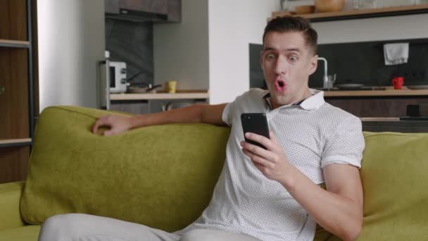 Şaşırtıcı mutlu genç adam başarıyı kutluyor akıllı telefona bakıyor ve evde kanepede otururken evet jestleri yapıyor. Mobil zafer kavramı — Stok video