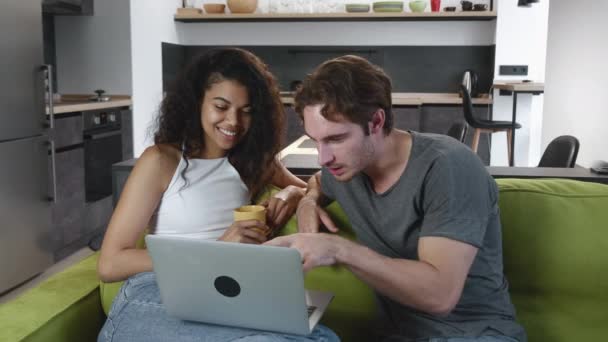 Sonriendo joven pareja multirracial disfrutando de discutir y usar el ordenador portátil mientras están sentados en el sofá en casa juntos, hombre y mujer felices mirando la diversión de la pantalla del ordenador portátil. — Vídeos de Stock