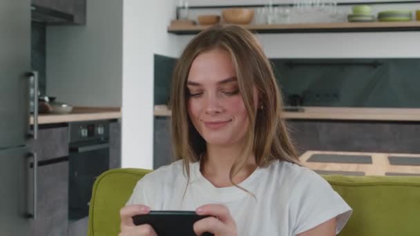 Szerencsejáték fiatal nő fehér pólóban játszani mobil játék mobiltelefon otthon. Vonzó mosolygós nő élvezi smartphone videojátékok ül kanapén a nappaliban. — Stock videók