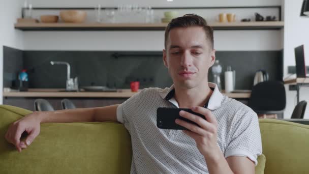 Joven alegre sentado en el sofá viendo videos divertidos en su teléfono inteligente sonrisa en el apartamento moderno — Vídeo de stock