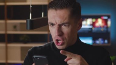 Akıllı telefon kullanan agresif bir adam. Adam modern bir evde otururken cep telefonundan okuduğu mesaja kızgın. Öfkeli erkek bağırır ve küfreder.