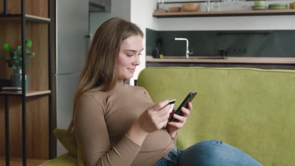 Felice giovane donna shopping online mentre riposava sul divano di casa. Donna sorridente online banking utilizzando smartphone shopping online con carta di credito in soggiorno. — Video Stock