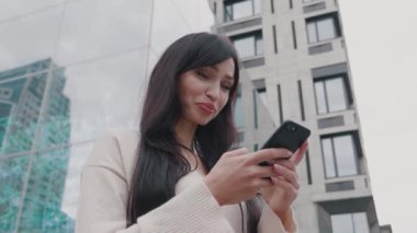 Gülümseyen iş kadını, açık havada, büyük modern binanın yakınında akıllı telefondan mesaj atıyor. Kentin arka planında cep telefonu uygulaması kullanan genç bir kadın.