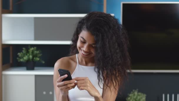 Ung afrikansk kvinde, der bruger smartphone surfing sociale medier, tjekker nyheder, sms 'er og smiler. Sød glad sort pige med krøllet hår ved hjælp af mobiltelefon derhjemme eller moderne kontor. – Stock-video