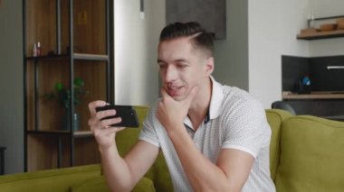 Kanepede oturup modern apartmandaki akıllı telefondan video izleyen genç adama yakın çekim.