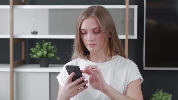 Besorgte Frau liest im Home Office schlechte Nachrichten auf ihrem Smartphone vor. Schockierte junge Frau hält Handy in den Händen — Stockvideo