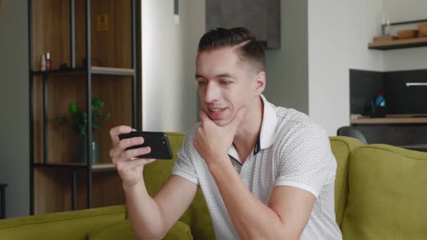 Primer plano del joven descansando sentado en el sofá viendo vídeo en el teléfono inteligente en el apartamento moderno — Vídeo de stock