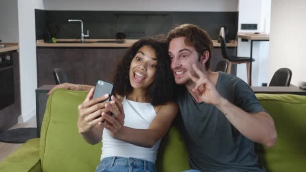 Funny interracial couple relaxing on couch taking selfie pictures using smartphone, enjoy weekend together posing for selfie on mobile phone for social media. — Stock Video