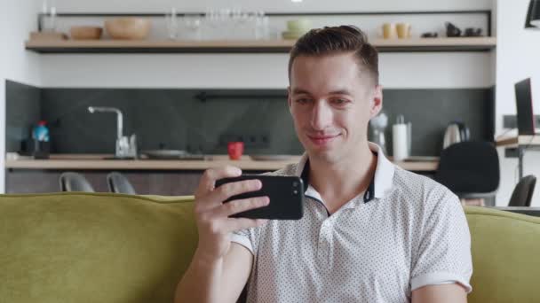 Retrato de un joven alegre sentado en el sofá viendo videos divertidos en su teléfono inteligente sonrisa en el apartamento moderno — Vídeos de Stock