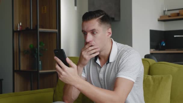 Shocked Man Mensaje de reacción en el teléfono inteligente se cubre la boca de susto sentado en el sofá en la sala de estar en casa. Desagradablemente sorprendido y asustado. Hombre se sorprende por lo que vio en su. — Vídeo de stock