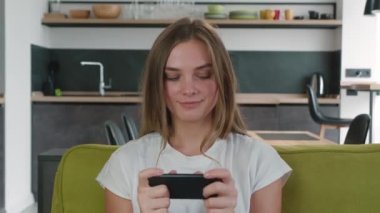 Beyaz tişörtlü genç kumarbaz kadın evde cep telefonuyla oyun oynuyor. Çekici, gülümseyen bir kadın oturma odasındaki koltukta oturmuş akıllı telefon video oyunlarından zevk alıyor..